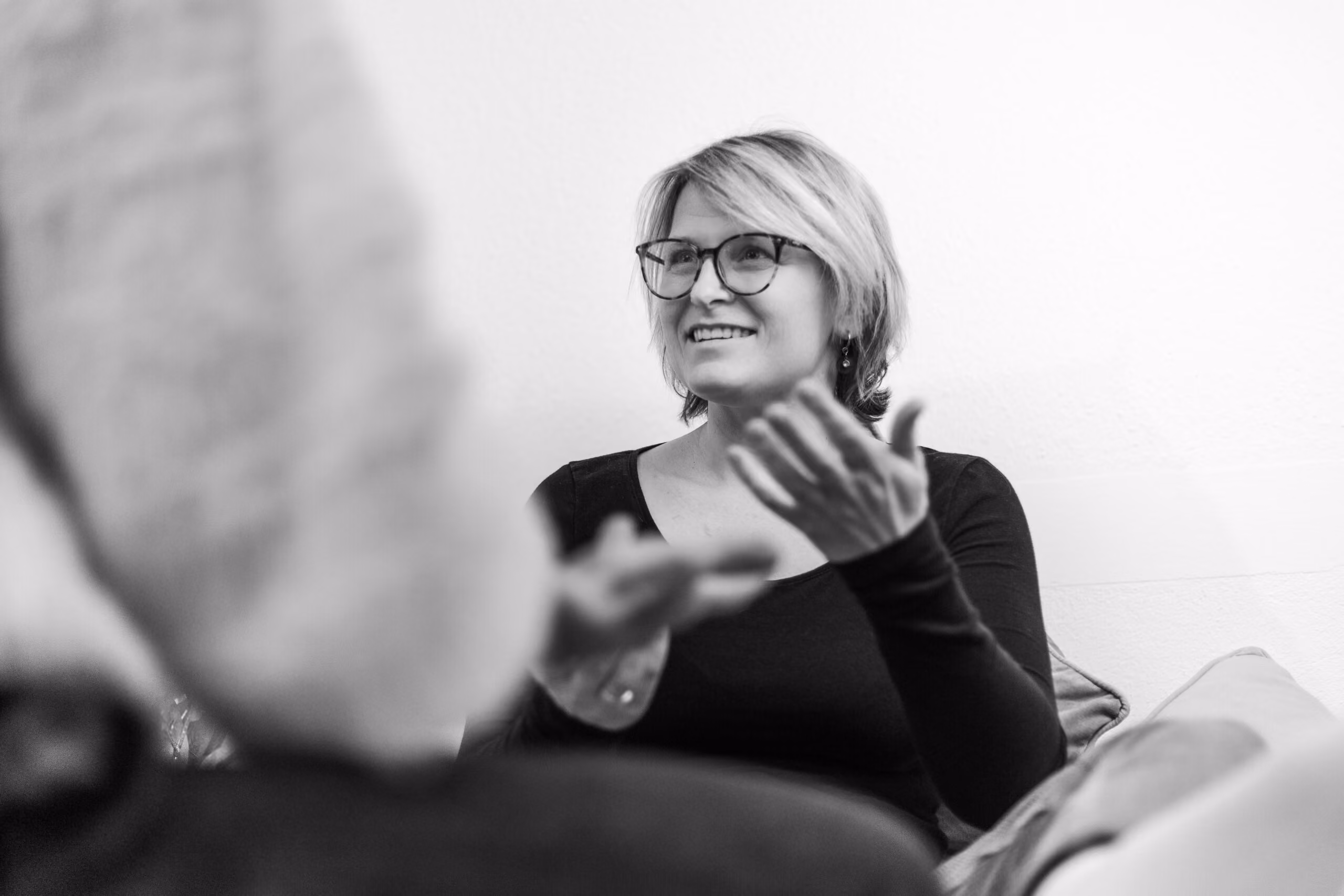 Schwarzweißfoto einer lächelnden Frau mit Brille, die in einem Gespräch mit einer anderen Person gestikuliert.