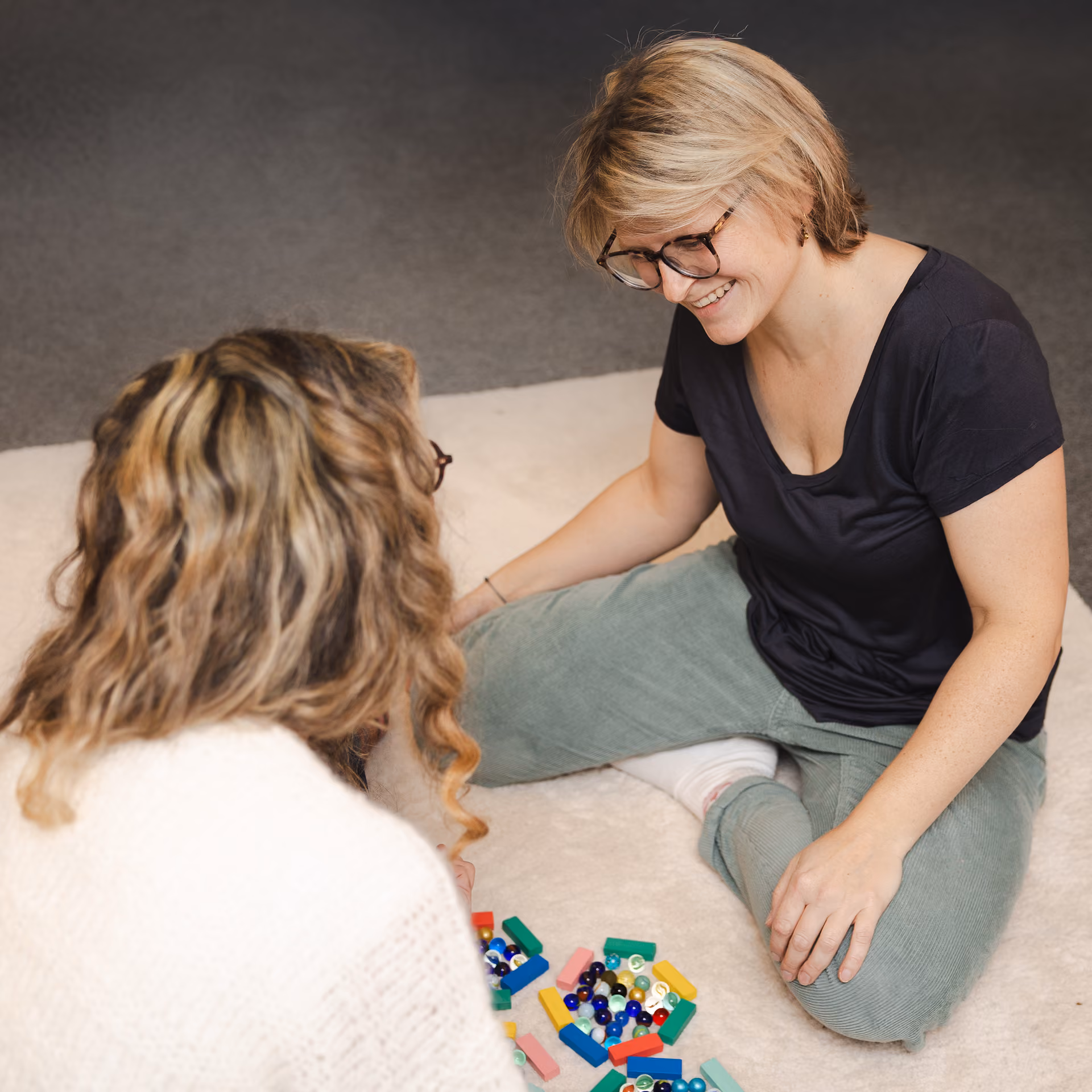 Zwei Frauen sitzen auf einem hellen Teppich und spielen mit bunten Murmeln und Bausteinen, eine Frau lächelt dabei.