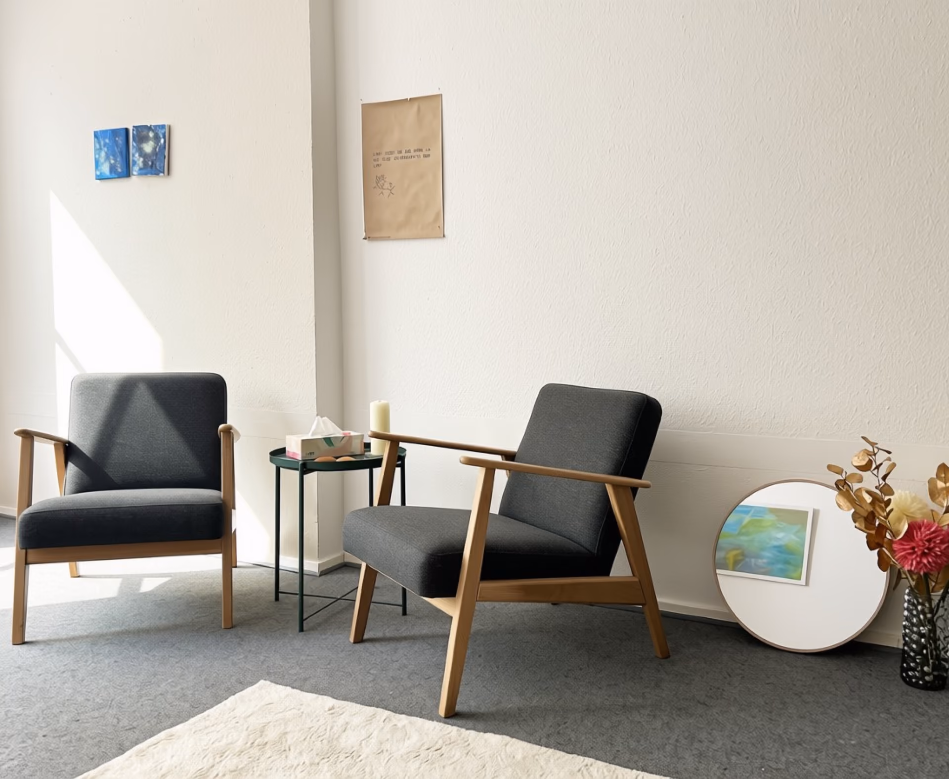 Zwei moderne graue Sessel mit Holzrahmen stehen in einem hellen Raum auf grauem Teppichboden, dazwischen ein kleiner runder Beistelltisch mit einer Kerze und einer Tissue-Box. An der Wand hängen zwei kleine blaue Bilder und ein braunes Papier mit Text, und eine runde Bodenleuchte spiegelt ein abstraktes Bild wider. Rechts steht eine Vase mit getrockneten Blumen.