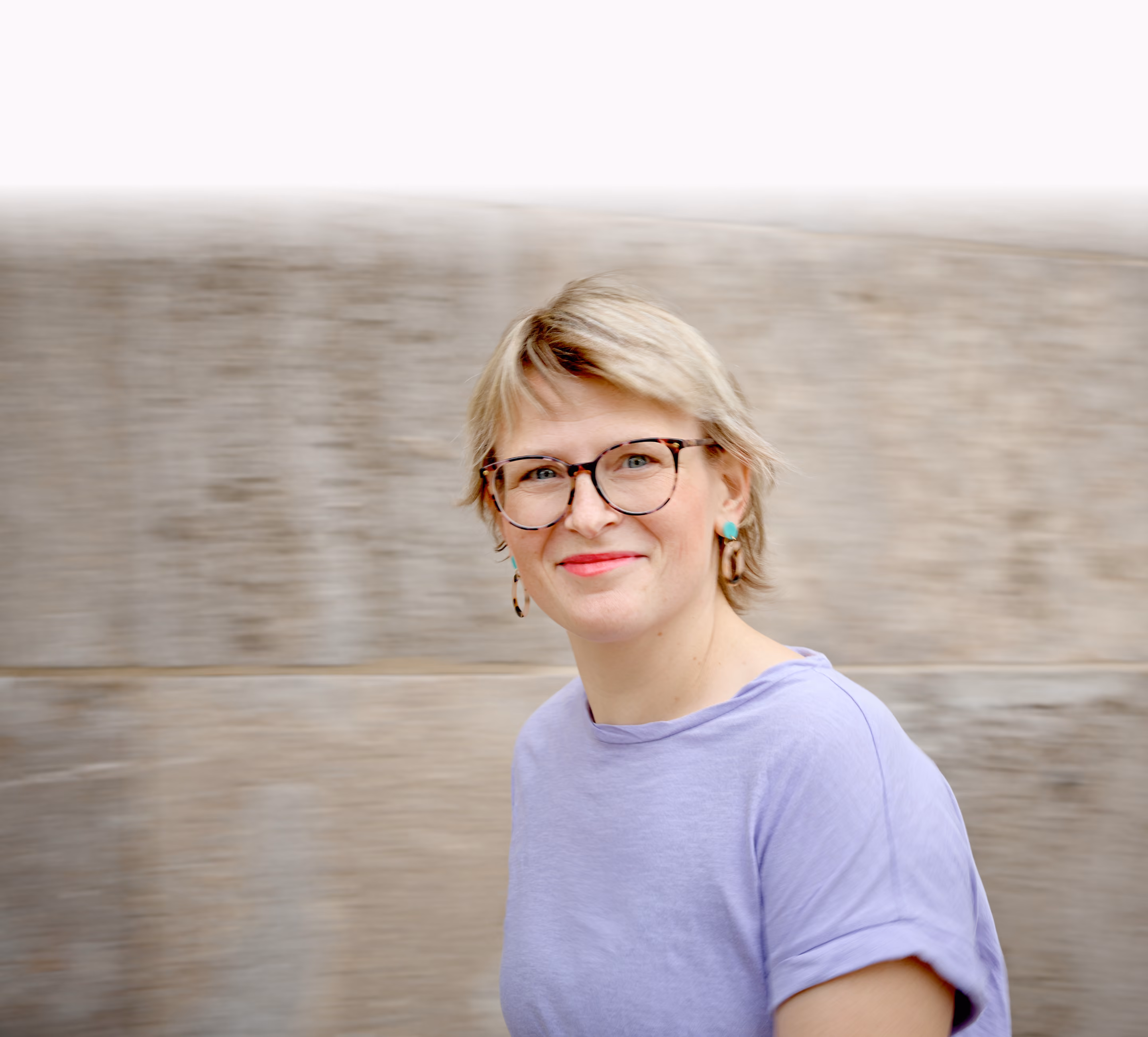 Frau mit kurzen blonden Haaren, die eine violette kurzärmelige Bluse, auffällige Ohrringe und eine Brille mit braunem Gestell trägt, lächelt vor einer unscharfen Steinwand.