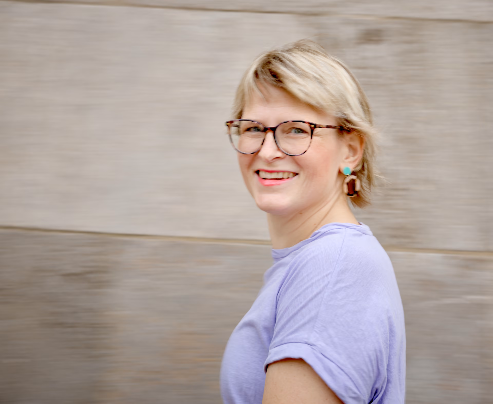 Frau mit kurzem blondem Haar, Brille und lila T-Shirt schaut lächelnd zur Seite vor einer grauen Steinwand.