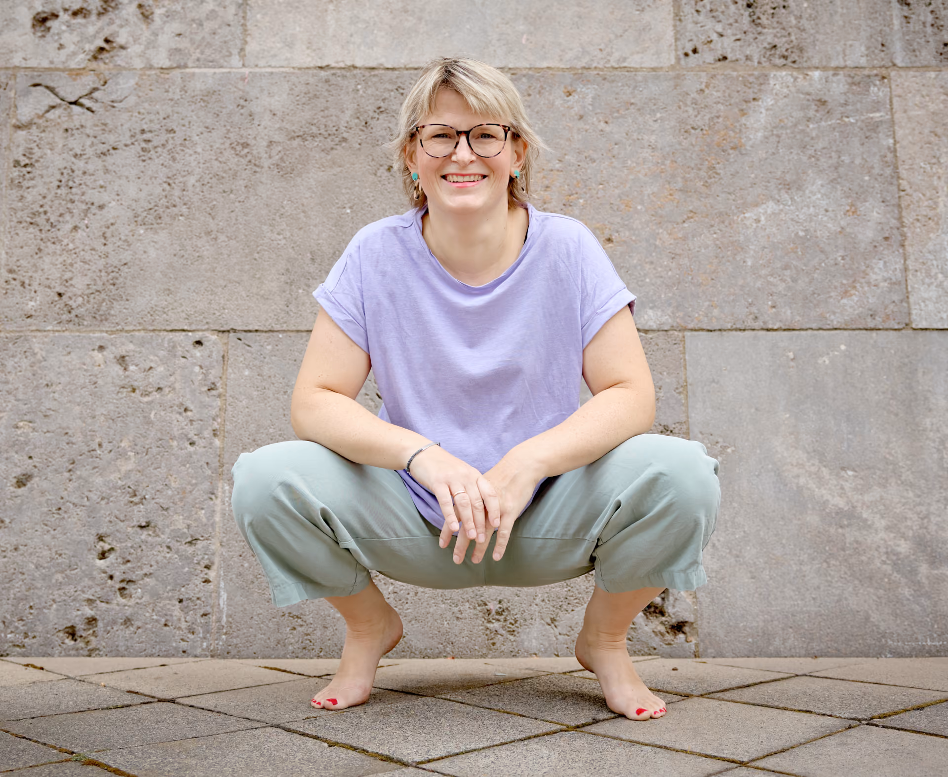 Frau mit kurzem blondem Haar, die eine lilafarbene Bluse und grüne Hose trägt, barfuß in einer Hockposition vor einer Steinfassade lächelnd in die Kamera schaut.