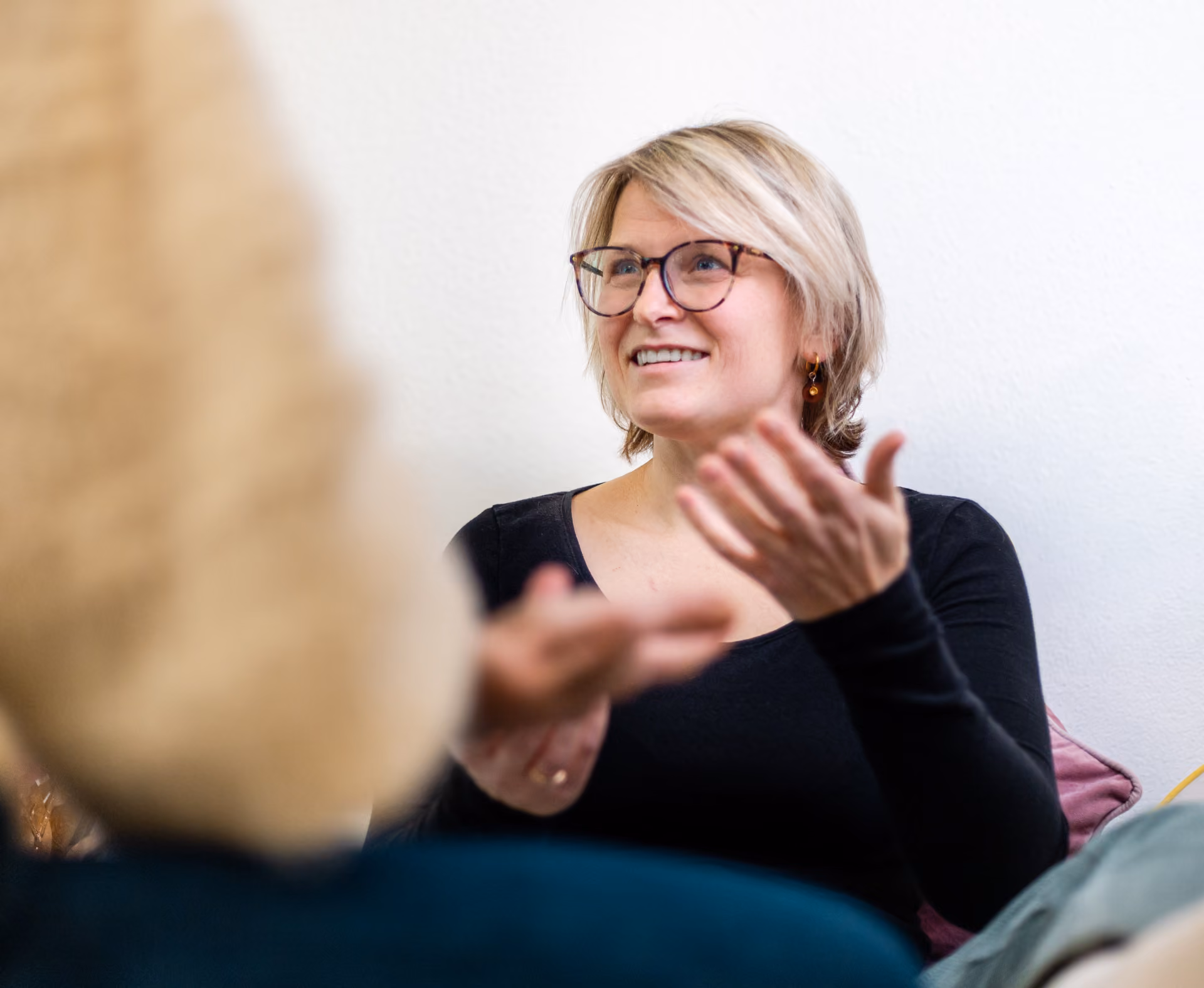 Frau mit kurzen blonden Haaren und Brille, die lächelnd und gestikulierend mit einer anderen Person spricht.