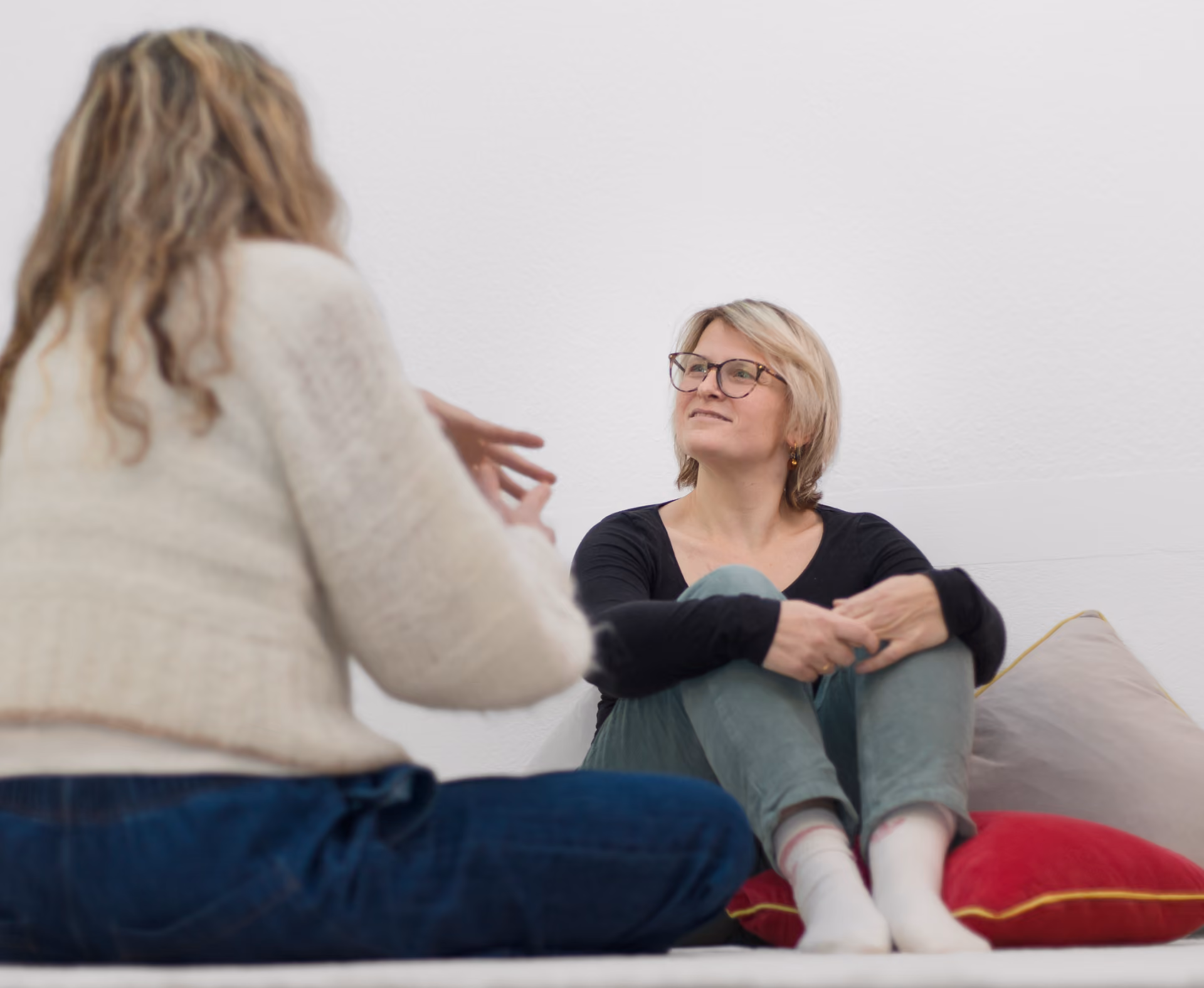 Zwei Frauen sitzen auf dem Boden in einem Gespräch, eine schaut aufmerksam zur anderen, die gestikuliert.