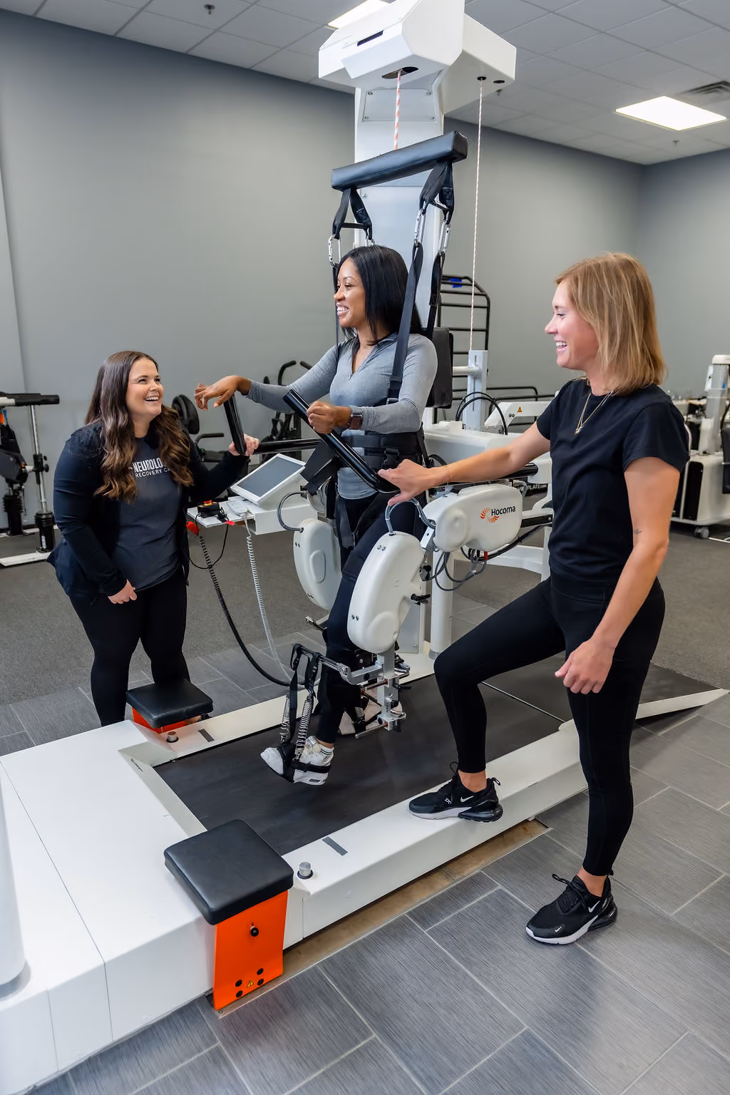 Two NeuroBridge clinicians smiling with a patient during a robotic gait-training session