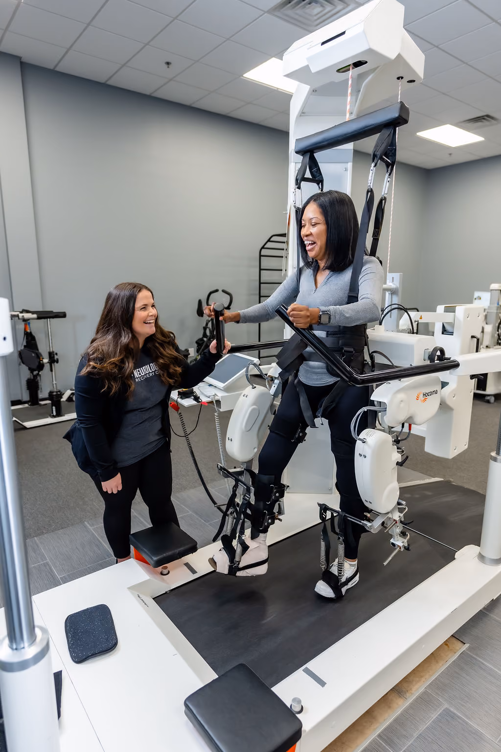 A NeuroBridge clinician supporting a smiling patient on a robotic gait trainer