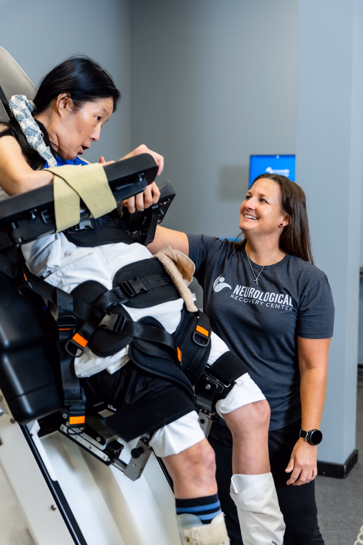 A NeuroBridge clinician encourages a patient strapped into a robotic tilt-table stepper