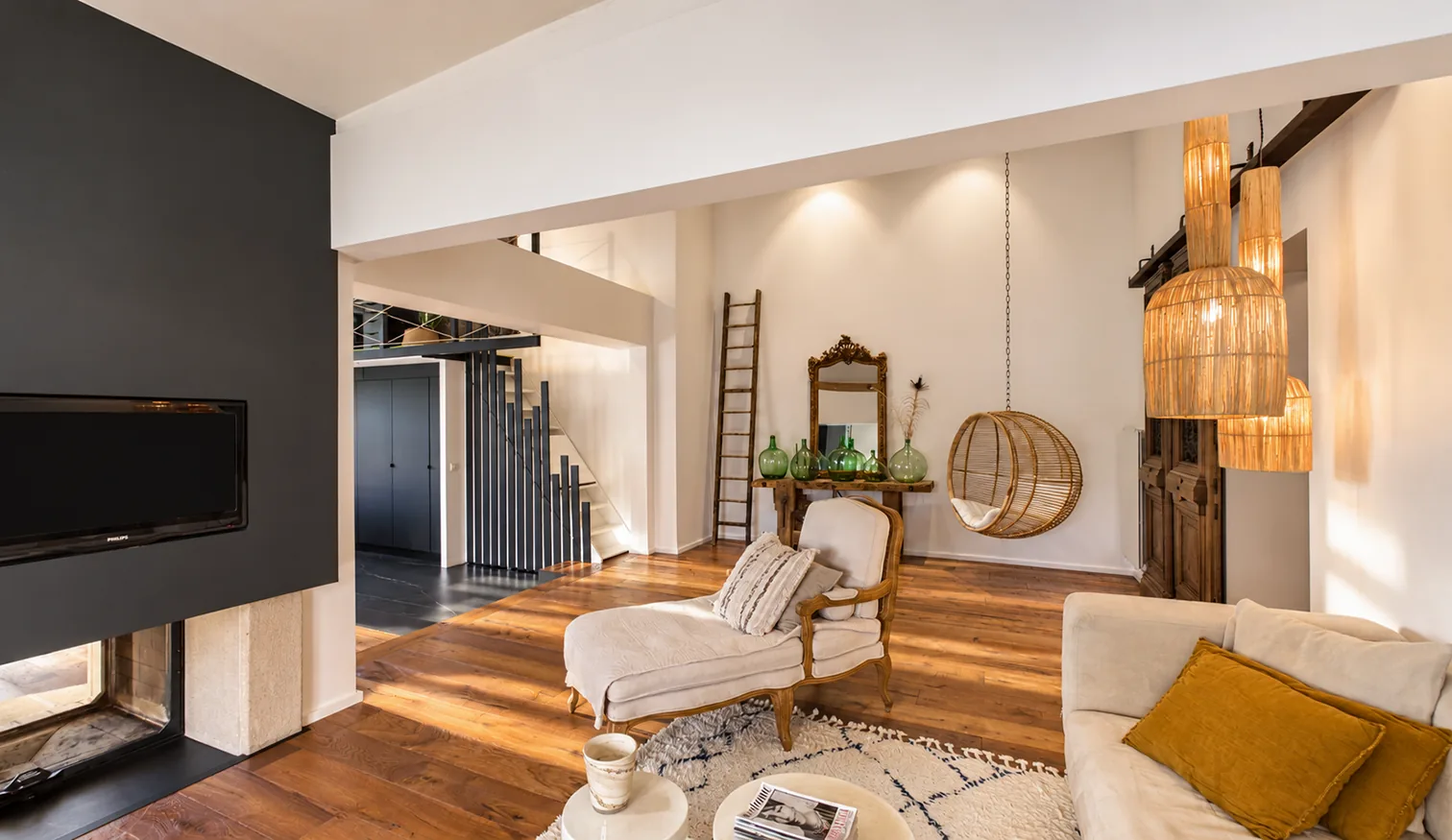 Salon lumineux avec parquet en bois, fauteuils clairs, suspensions en osier et cheminée noire.