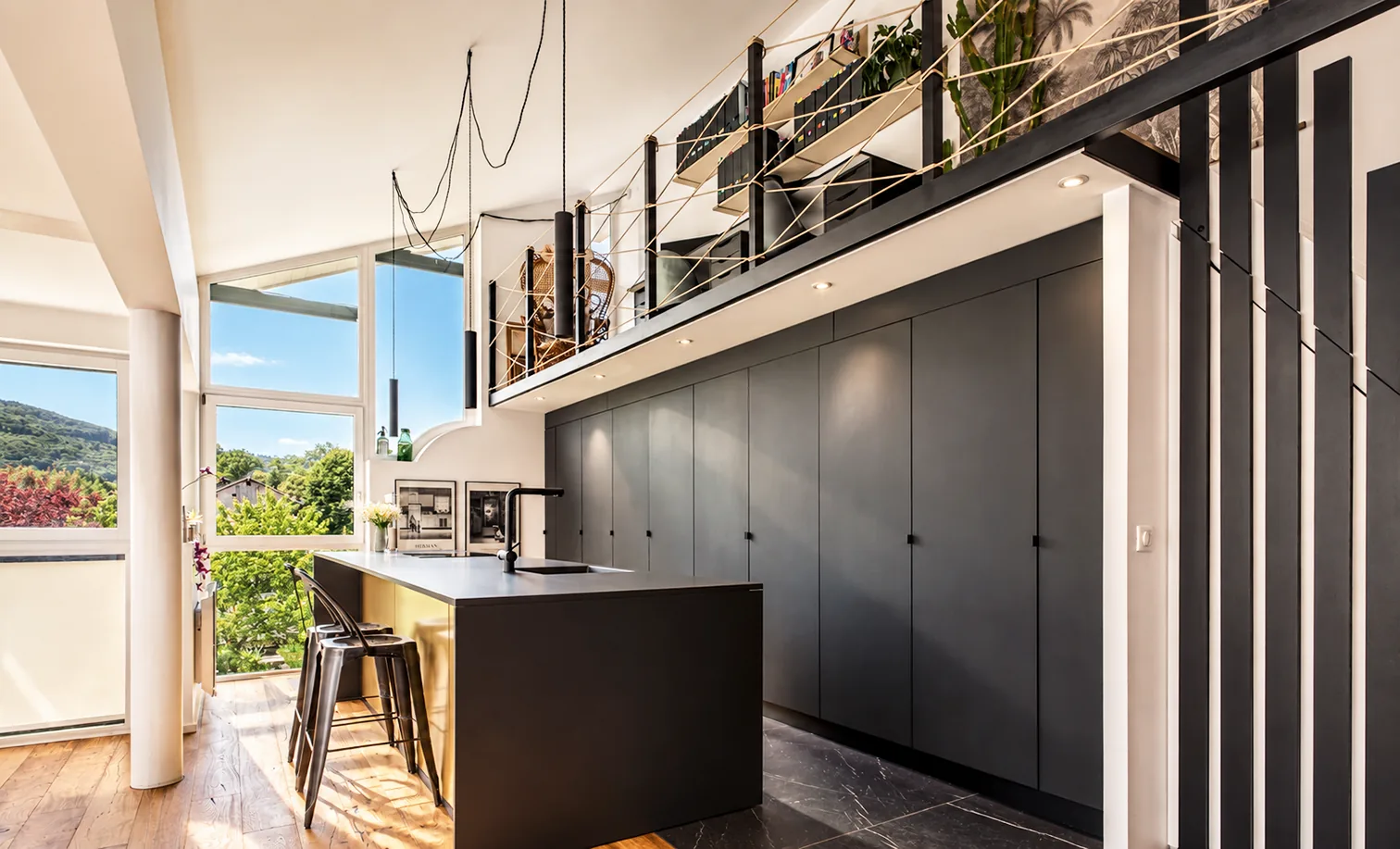 Cuisine avec îlot noir et mur de rangements noirs, baie vitrée sur la verdure et parquet en bois.