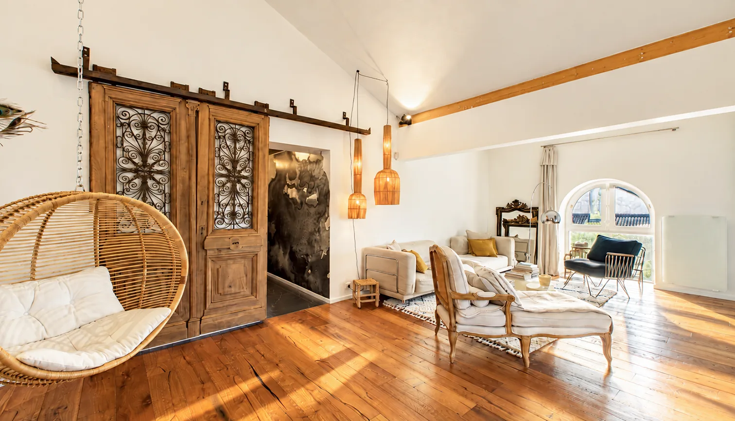 Salon rénové avec parquet en bois, portes anciennes coulissantes, fauteuil suspendu et baie cintrée.