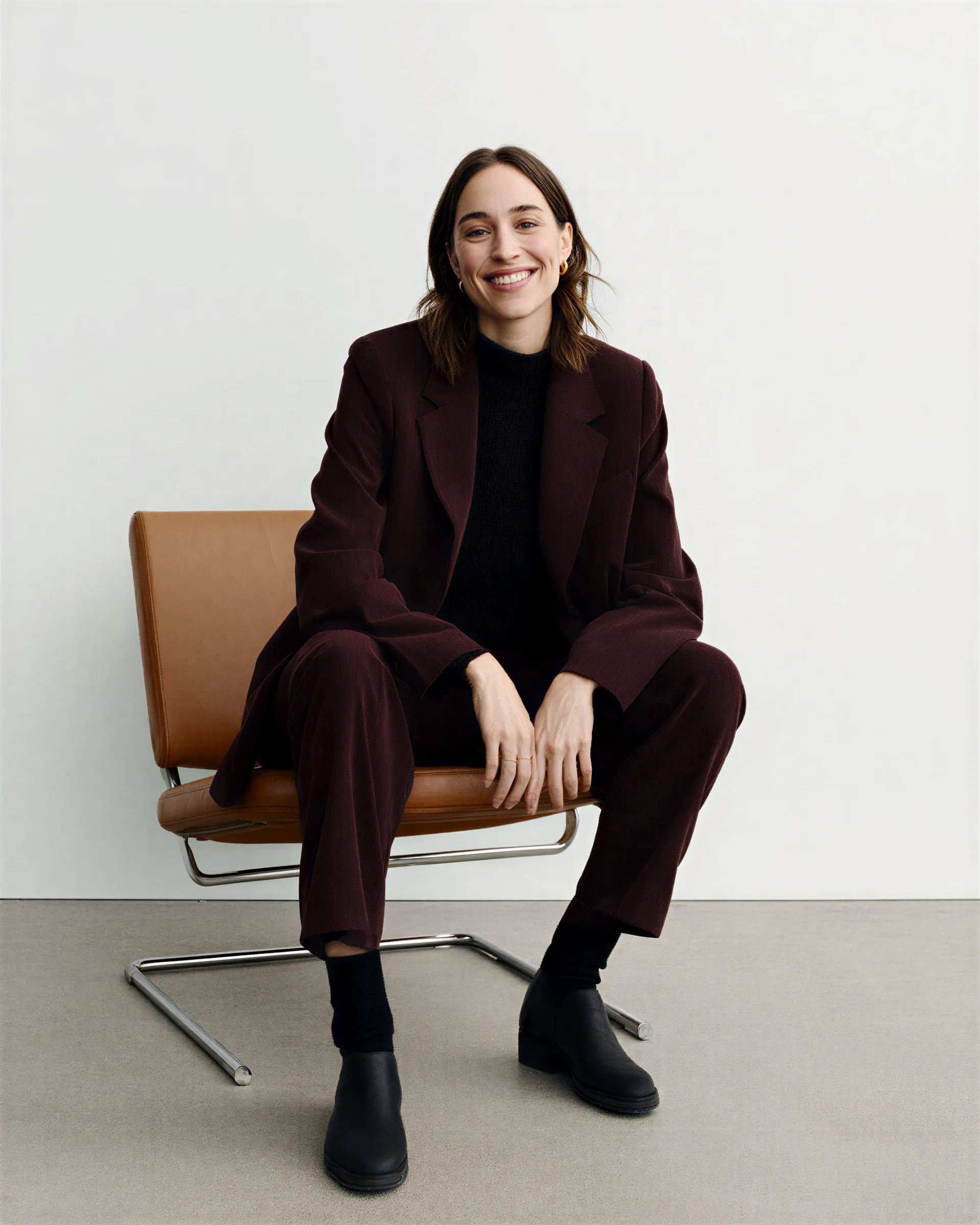 Smiling woman wearing a dark suit sitting on a brown leather chair against a plain white wall.