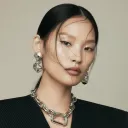 Portrait of a woman with smooth dark hair styled back, wearing large chain-link earrings and matching necklace against a plain background.