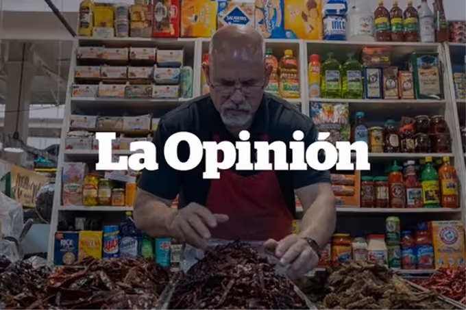 A man wearing glasses and a red apron arranges piles of dried chili peppers at a market stall with shelves of various packaged goods behind him.