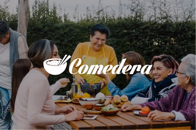 A group of people around a wooden outdoor table sharing a meal and enjoying each other's company, with a woman in a yellow apron preparing food in the center and the word 'Comedera' overlayed.