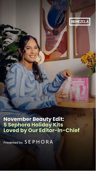 Smiling woman with long dark hair in a blue and gray sweater sitting at a wooden table holding a pink Sephora beauty kit product, with two pink beauty kit packages on the table next to a vase of yellow flowers and framed abstract art on the wall behind her.