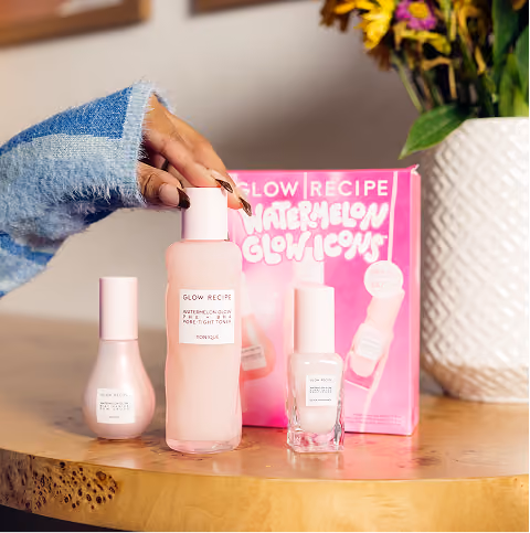 Hand with dark nail polish and a denim sleeve holding a pink bottle of Glow Recipe Watermelon Glow Pore Tight Toner on a wooden table next to two other pink Glow Recipe skincare bottles, with a pink product box and a white textured vase with yellow and purple flowers in the background.