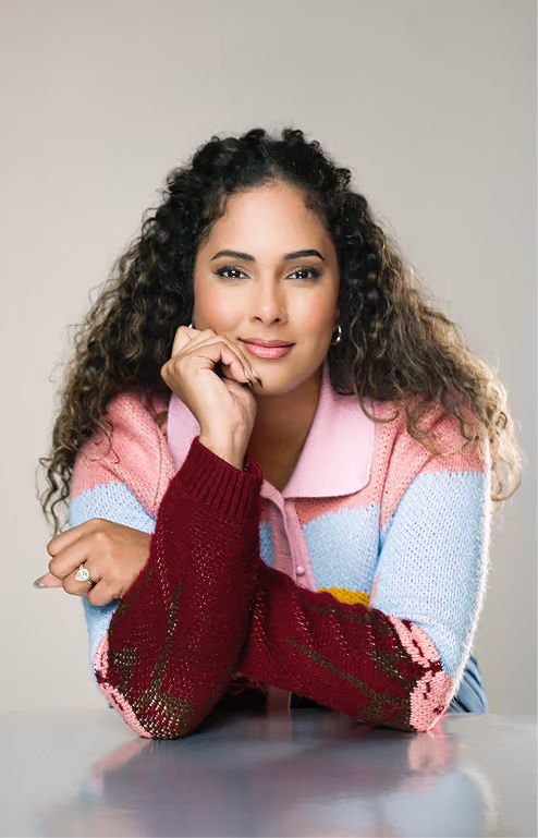 Woman with long curly hair resting her chin on her hand, wearing a colorful knitted sweater with pink, blue, yellow, and burgundy stripes, smiling gently in front of a neutral background.