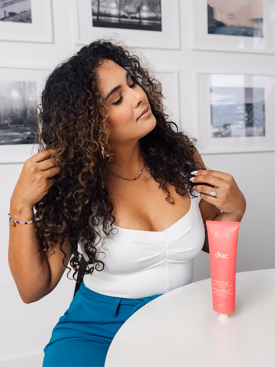 Woman with long curly hair sitting at a white table, touching her hair, with a pink tube of Dae hair product on the table in front of her, white wall with framed black-and-white photos in the background.
