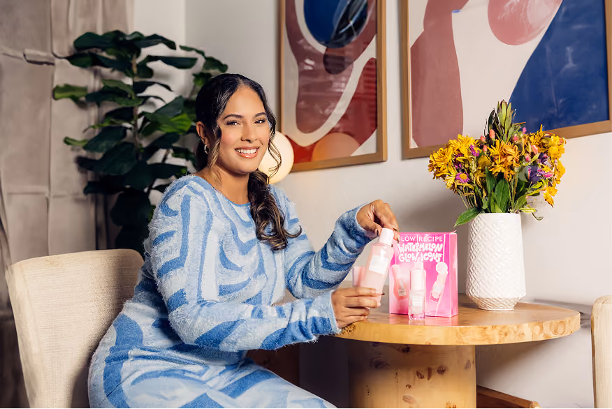 A smiling woman in a blue and white patterned sweater and skirt sits at a round wooden table holding skincare products, with a pink product box and a white vase with yellow and purple flowers on the table, and two abstract framed artworks on the wall behind her.