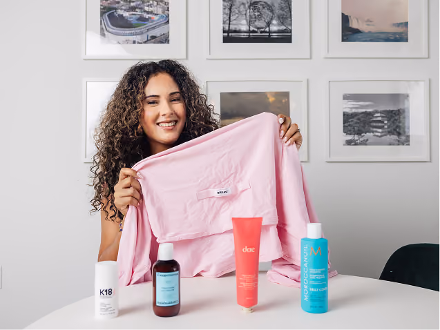 Smiling woman with curly hair holding a pink cloth labeled 'BRAVO' behind a white table displaying four hair care products in bottles and tubes.
