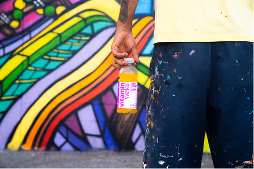 Person wearing paint-splattered dark shorts and a light shirt holding a bottle of Vitamin Water with a colorful graffiti wall in the background.