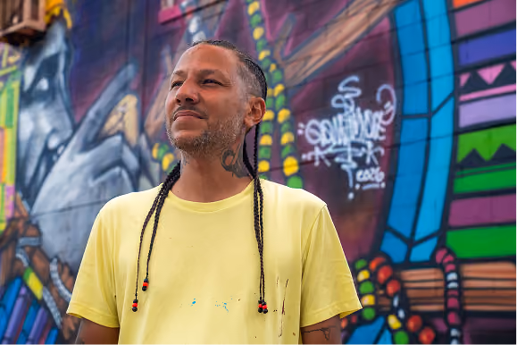 Man with braided hair wearing a yellow t-shirt standing in front of a colorful graffiti wall with abstract and geometric patterns.