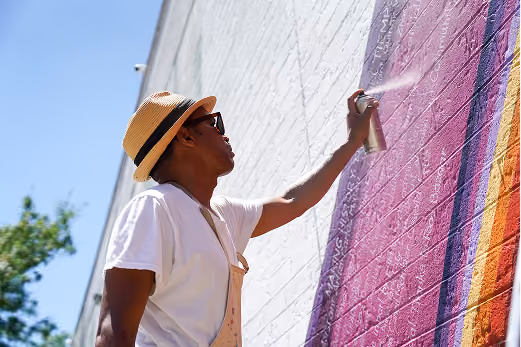 Person wearing a white T-shirt, sunglasses, and a straw hat spray painting a colorful mural on a large white brick wall outdoors.