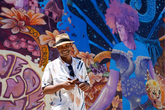 Smiling man wearing a hat and patterned shirt holding a phone, standing in front of a vibrant mural featuring a large purple-haired figure, flowers, and colorful graffiti elements.