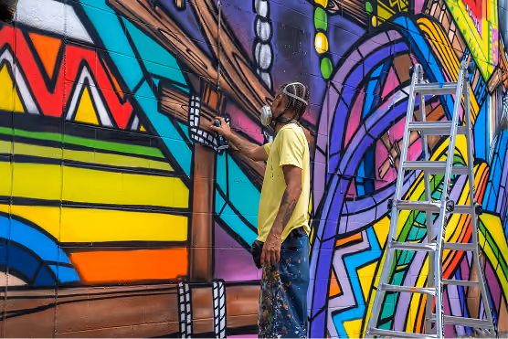 Man wearing a yellow shirt and face mask spray painting a colorful geometric mural on a brick wall, standing beside a metal ladder.