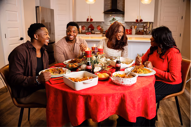 Four adults happily sharing a meal around a round table covered with a red tablecloth, featuring dishes of food and two bottles of Coca-Cola in a warmly lit kitchen decorated for Christmas.