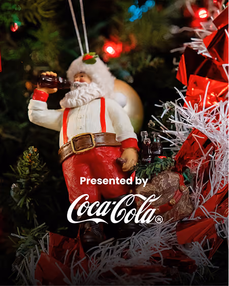 Christmas ornament of Santa Claus with white beard, red suspenders, and red pants holding and drinking from a small Coca-Cola bottle, hanging on a decorated Christmas tree with red and white ribbons and lights.