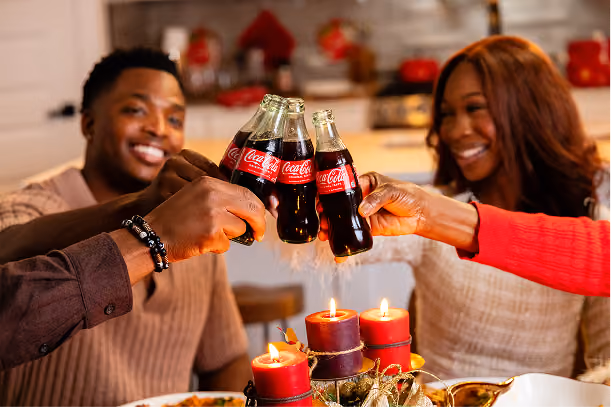 Four people smiling and toasting with glass Coca-Cola bottles over a table with lit red candles and festive decorations in a warmly lit room.