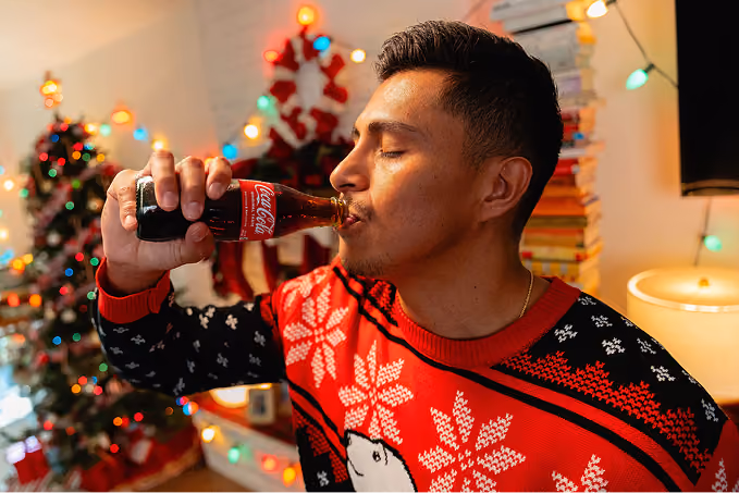 Man wearing a red and black Christmas sweater with white snowflakes drinking from a glass bottle of Coca-Cola in a room decorated with colorful holiday lights and Christmas trees.