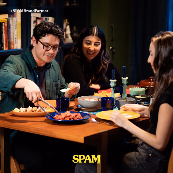 Three people sitting around a wooden table sharing a meal featuring SPAM, with one person serving pieces onto a tortilla while others smile and engage in conversation, set in a warm indoor environment.