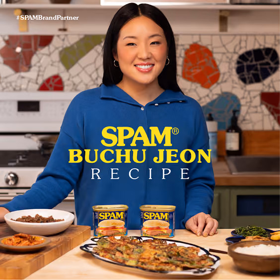 Smiling woman in a blue sweater stands behind a kitchen counter with two cans of SPAM, a plate of buchujeon (Korean savory pancakes), and bowls of side dishes, with a tiled wall in the background and text 'SPAM Buchu Jeon Recipe'.