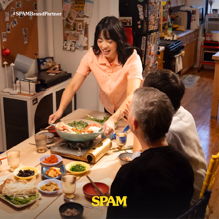 A woman in a peach shirt serving a steaming hotpot meal to two seated men at a dining table filled with dishes of vegetables and side dishes in a cozy kitchen setting.