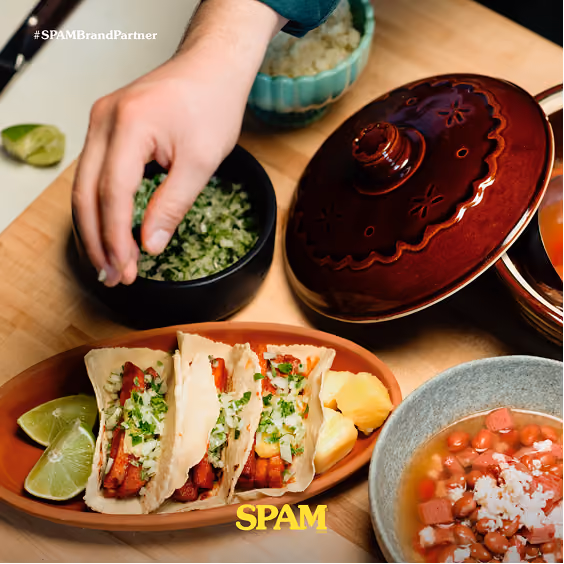 Three tacos filled with grilled meat, chopped onions, and cilantro on an oval plate with lime wedges and pineapple chunks, next to a bowl of beans topped with cheese and a hand reaching toward a bowl of chopped green herbs.