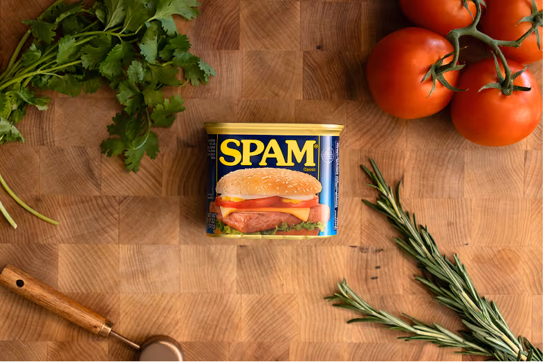 A can of SPAM Classic placed on a wooden cutting board surrounded by fresh herbs including cilantro and rosemary, a cluster of vine tomatoes, and a wooden-handled kitchen tool.