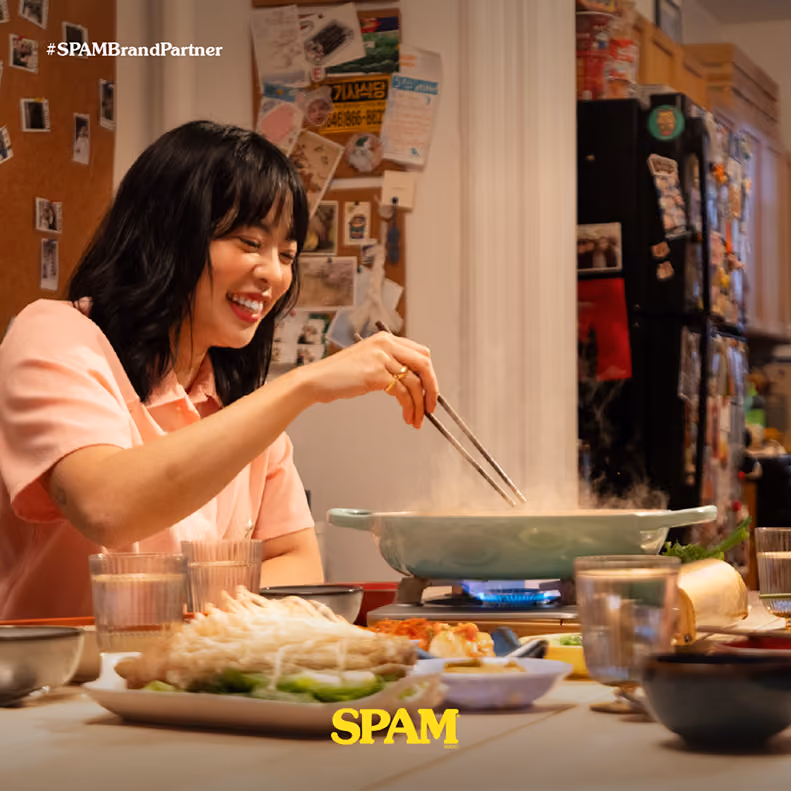 Smiling woman using chopsticks to cook in a steaming pan on a portable stove at a table set with various dishes and glasses in a cozy kitchen.