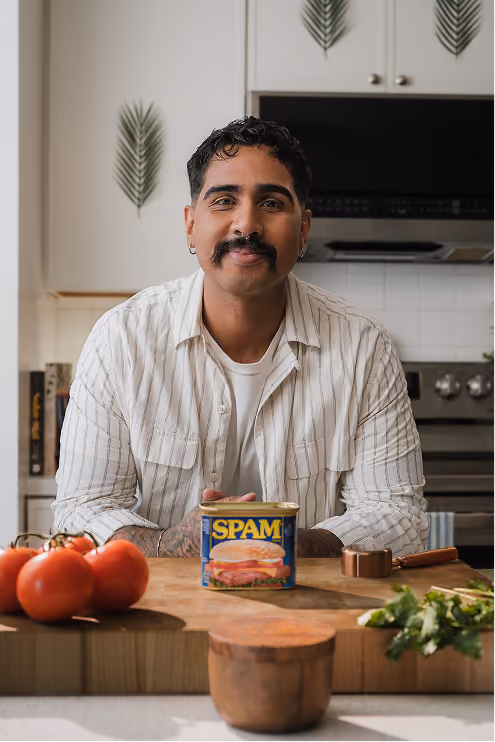 Man with dark hair, mustache, and earrings smiling in a kitchen, leaning on a wooden counter with a can of SPAM, tomatoes, and greens in front.