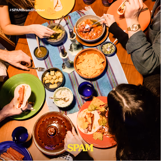 Overhead view of a table with four people eating a Mexican-style meal featuring tortillas with hot dogs, bowls of salsa, guacamole, cheese cubes, and tortilla chips on colorful plates and a striped table runner.