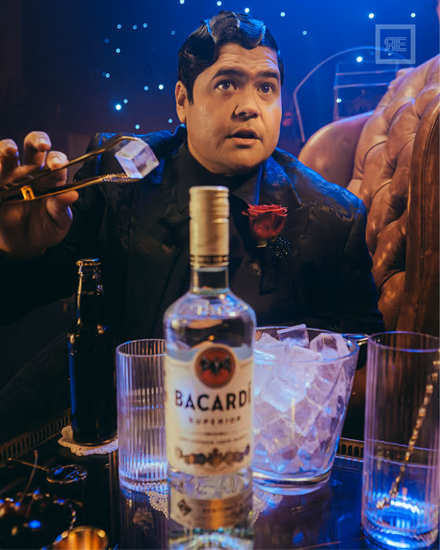 Man with styled hair and a red rose on his black suit adding ice cubes with tongs to a glass, seated near a table with a bottle of Bacardi rum, a bucket of ice, and two empty glasses in a dimly lit room with blue lights.