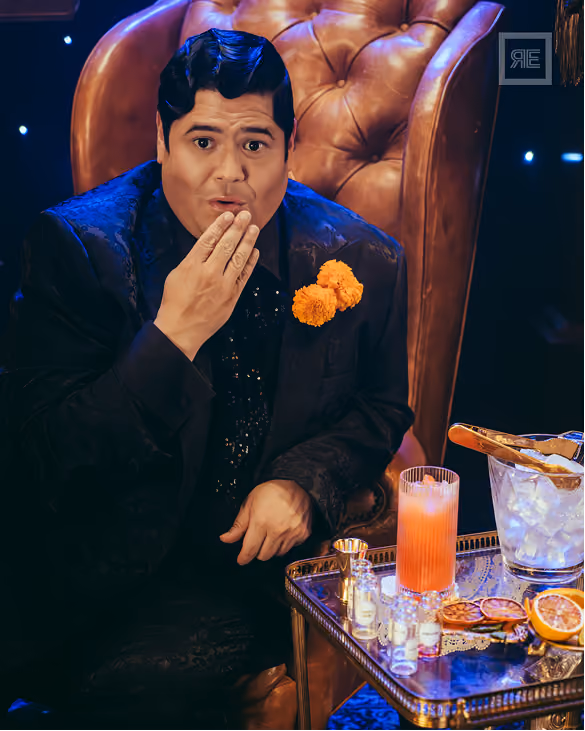 Man in a black suit with orange flowers on the lapel sitting on a brown leather chair, covering part of his mouth with his hand, next to a glass table holding a tall pink drink, an ice bucket with tongs, and garnished dried citrus slices.