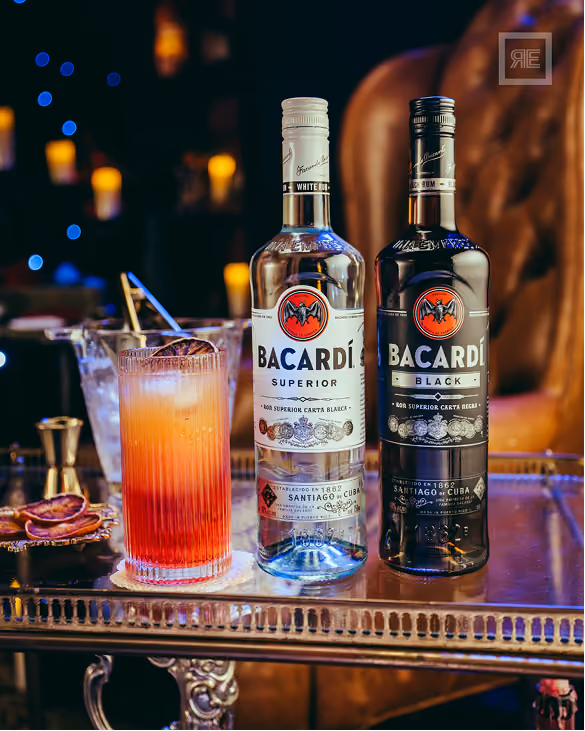 Two Bacardi rum bottles, Bacardi Superior with white label and Bacardi Black with black label, placed side by side on a reflective metal table next to a tall glass with an orange to red gradient cocktail and a metal straw, with a dimly lit background featuring blue and yellow blurred lights.
