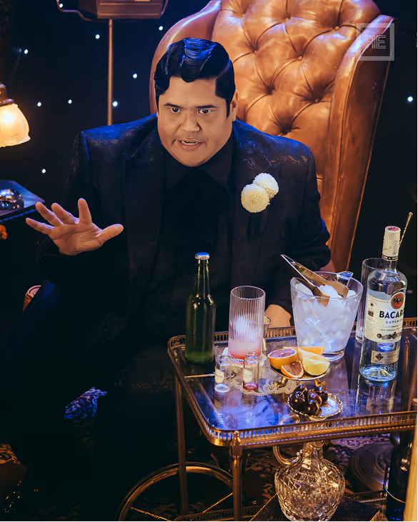 Man dressed in black suit with white flower on lapel seated in a large brown leather armchair gesturing with his right hand, in front of a glass table holding a green bottle, a glass with a pink drink, an ice bucket, a bottle of Bacardi rum, sliced citrus fruits, and a small plate of cherries.
