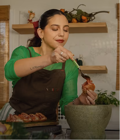 A woman with dark hair tied back, wearing a green shirt and brown apron, is holding a piece of food and using a spoon to add sauce or topping inside it in a kitchen setting with plants and shelves in the background.