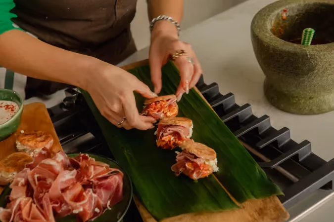 Person assembling small sandwiches or sliders with crispy crust, wrapped with thin slices of cured meat on a green banana leaf on a stove, with a separate plate of cured meat and a green stone mortar with pestle in the background.