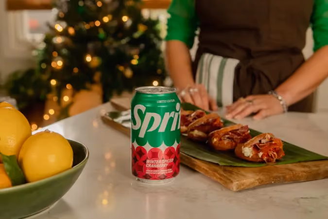 A limited edition green can of Sprite Winter-Spiced Cranberry placed on a kitchen counter with a person in the background preparing wrapped snacks on a wooden cutting board, and a bowl of lemons on the left side.