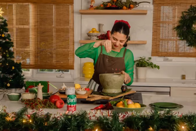 Woman in a green shirt and brown apron uses a large mortar and pestle in a kitchen decorated with Christmas garlands, a tree, and a wreath.
