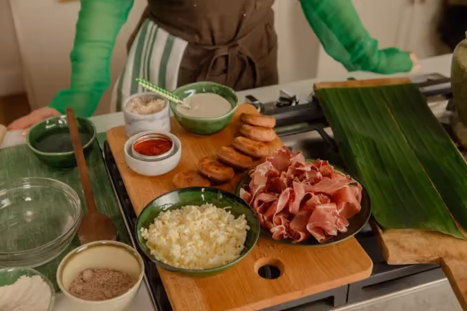 Wooden cutting board on a kitchen stove with ingredients including sliced plantains, shredded cheese, red sauce, a brown powdered spice, and thinly sliced cured meat, with a person in green sleeves standing behind.